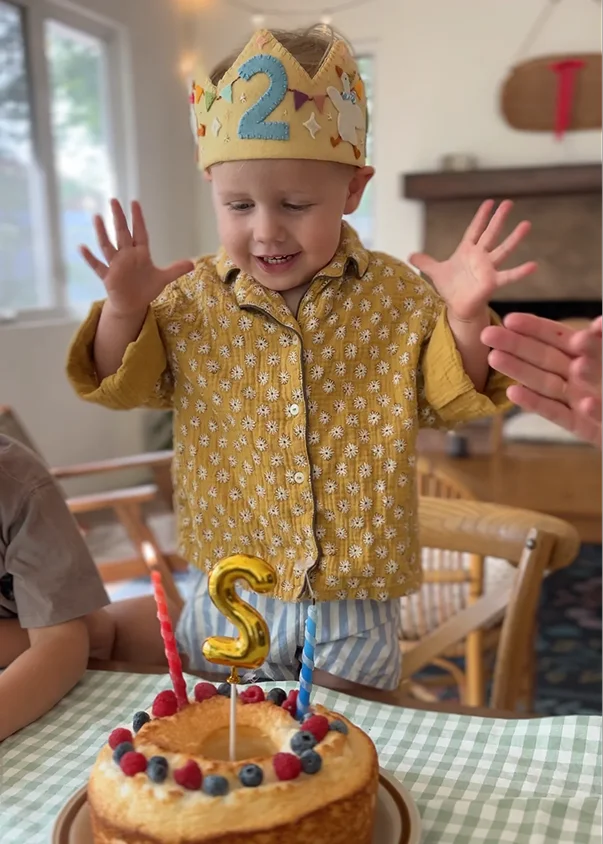 Menino de 2 anos usando a coroa de feltro com o número 2 na frente, diante do bolo de aniversário